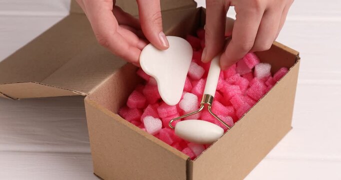 Woman putting styrofoam pieces, face roller and gua sha tool into cardboard box at white wooden table, closeup