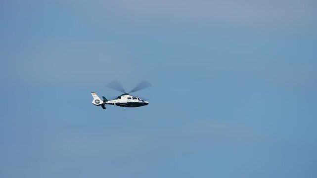 White twin-engine helicopter with green accent livery in steady forward flight against clear blue sky with rotors spinning