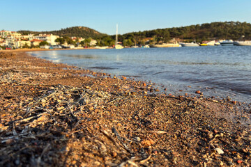 Fototapeta premium Scenic Turkish Aegean coast with pebble beach, calm blue sea, and boats moored near tranquil seaside resort town on a sunny day; ideal for summer travel, Mediterranean vacation, and tourism concept