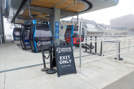 Grouse Mountain branded EXIT ONLY DO NOT ENTER sandwich board sign at gondola station with cabins and chalet buildings behind on a foggy spring day.