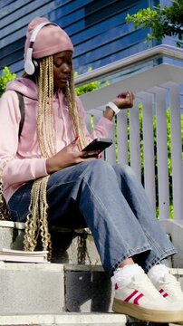 Vertical video: Scrolling student in pink hoodie tapping phone on steps, listening with headphones