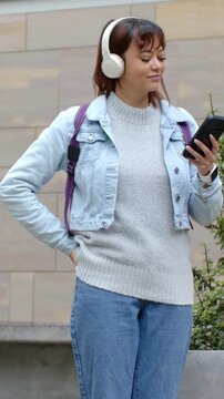 Vertical video: Glancing, woman with backpack and headphones replying to message on phone at plaza