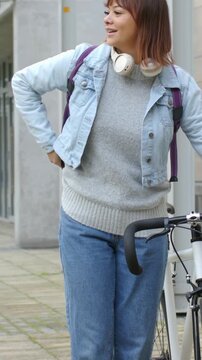 Vertical video: Adjusting backpack strap, woman in denim taking phone and calling by building, bike