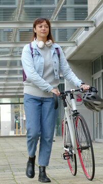 Vertical video: Starting under glass canopy, woman pushing bike toward lens in denim with backpack