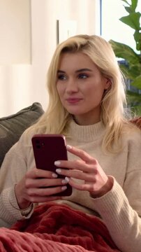 Vertical video: Phone buzzing, woman in knit holding red phone reading messages on gray couch