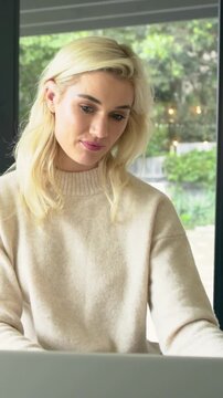 Vertical video: Gazing down at laptop, woman in tan top sipping mug, typing to focus by glass doors