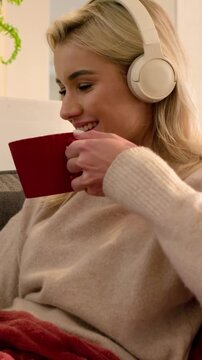 Vertical video: Settling woman with headset sipping red mug and checking tablet at home, copy space