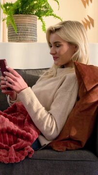 Vertical video: Reading message, woman speaking into smartphone on sofa in cream sweater and jeans
