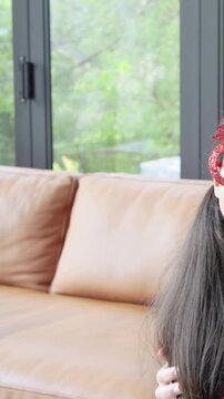 Vertical video: Smiling girl in tree-glasses on couch at home, woman Santa hat leaning causing pan