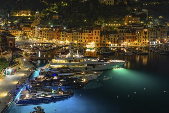 Luxury yachts and boats anchored in Portofino harbour at night in front of illuminated pastel-coloured house facades, view from San Giorgio church, Portofino, Liguria, Italy