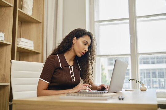 In a cozy home office, a woman with long, curly hair focuses intently on her laptop while seated at a wooden desk