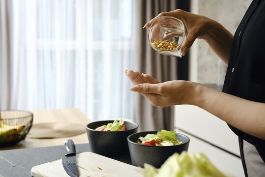 A person adds pine nuts to a bowl filled with fresh salad ingredients, including lettuce and tomatoes, in a modern kitchen