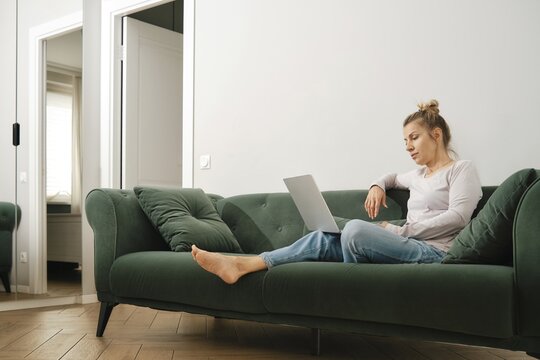 Middle-aged woman is sitting comfortably on a green sofa in a bright, modern living room, focused on her laptop. She is watching a subscription movie on an online streaming platform