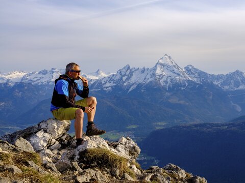Mountaineer with a snack at the summit Rauher Kopf, Berchtesgadener Alps, behind Watzmann, Bischofswiesen, Berchtesgadener Land, Upper Bavaria, Bavaria, Germany