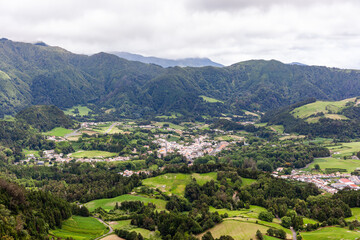Fototapeta premium Furnas village nestled in lush green valley with surrounding mountains. Scenic view from Pico do Ferro Viewpoint showcasing volcanic landscape.
