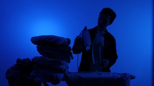 Man in silhouette ironing a stack of neatly folded clothes on an ironing board against a vibrant blue background, domestic routine and quiet household chore