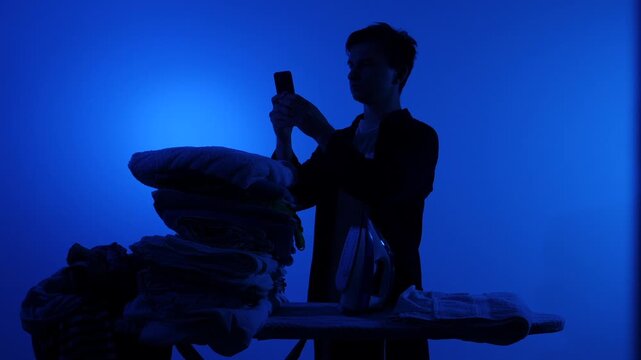 Man's silhouette at an ironing board, glued to his phone while an iron and pile of laundry are ignored against a vivid blue backlit background, sign of domestic distraction