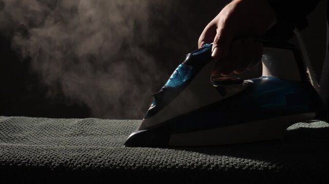 Hand held iron pressing garment with steam rising in dramatic silhouette against dark background, close up of domestic chore and fabric care showing heat, steam, and smoothening action