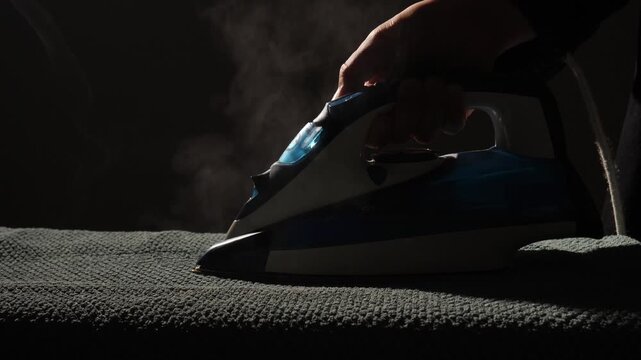 Person's hand powerfully operating a steam iron over fabric, creating a stream of vapor while performing domestic household chores in a dark, moody setting