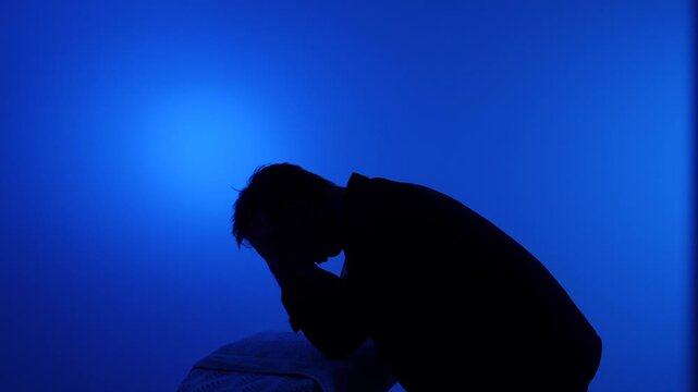 Man in silhouette covering his face with hands, feeling exhausted and frustrated while taking a break from ironing clothes at home against a deep blue background
