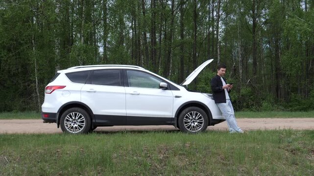 Young man standing beside broken white suv with hood open on rural road, using smartphone to call roadside assistance while waiting alone near forest, frustrated and worried