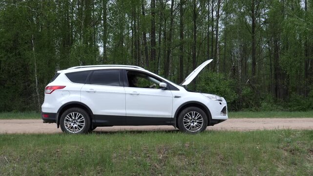 White suv experiencing mechanical breakdown on a rural dirt road, with the hood open, indicating engine trouble and needing assistance in a forest environment