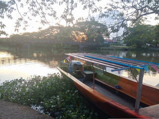 Naklejka premium A traditional wooden boat is moored along a calm riverbank during a peaceful sunset, featuring a distant truss bridge.