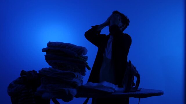 Man silhouette covering his face in exhaustion while ironing a towering stack of laundry on a blue background, conveying household overload, stress and domestic fatigue