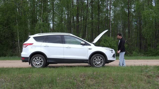 Man examining suv engine with hood open, facing a car breakdown problem on a remote dirt road next to a forest, representing vehicle issues and roadside assistance needs