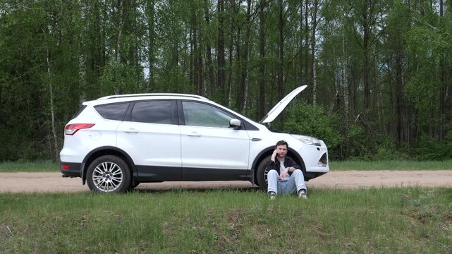 Young man sitting next to a white car with open hood on a dirt roadside, using a smartphone to call for assistance after a car breakdown in the forest