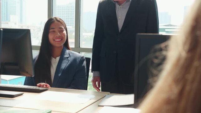 Asian manager behind young businesswoman working at computer, smiling colleagues looking at desktop monitor together, modern office workplace with city, senior boss providing supervision to employee.