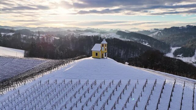 Aerial view of Dreisiebner Kapelle on a snow-covered hill surrounded by winter vineyards and rolling hills in Gamlitz, Styria, Austria.