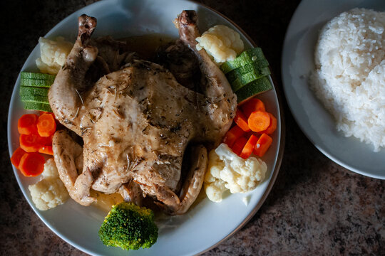 Pollo al horno a las yerbas con mantequilla servido en un plato blanco con vegetales cocidos como zanahoria en rodajas y trozos de coliflor, con arroz a vapor al lado, claroscuro o low key light