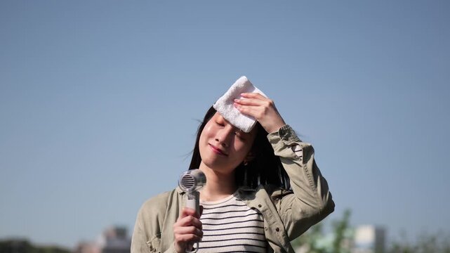 夏の青空の下でハンディファンとハンカチで汗を乾かす女性