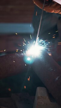 Closeup of skilled welder joining steel pipe with electric arc in dark workshop environment. Bright sparks and glowing metal create dramatic industrial scene focused on precision craftsmanship.