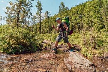 Obraz premium hiking teenager with a large backpack crossing a shallow mountain stream in a sunny green forest