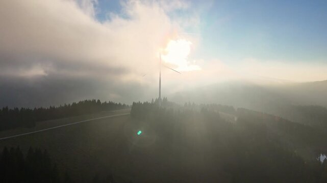 Aerial view of Windpark Freilanderalm wind turbine standing on a forested hill with sun rays piercing through thick fog and clouds in Modriach, Styria, Austria.