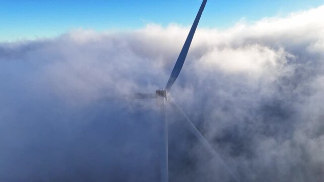 Aerial view of a wind turbine at Windpark Freilanderalm emerging from thick clouds under a clear blue sky in Modriach, Styria, Austria.