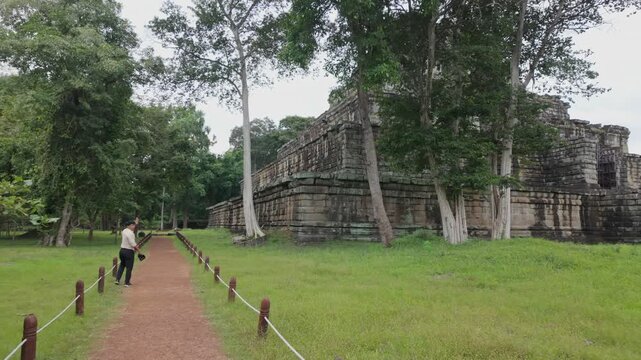 A lone man walks along a path towards ancient stone ruins in Koh Ker Pyramid Temple, Cambodia. The jungle setting highlights the historical significance of the site.
