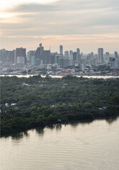Fototapeta premium A vast expanse of dense, emerald canopy blankets the peninsula of Bang Kachao, standing as a tranquil fortress of nature against the encroaching modern world. In the foreground, the glassy waters of t