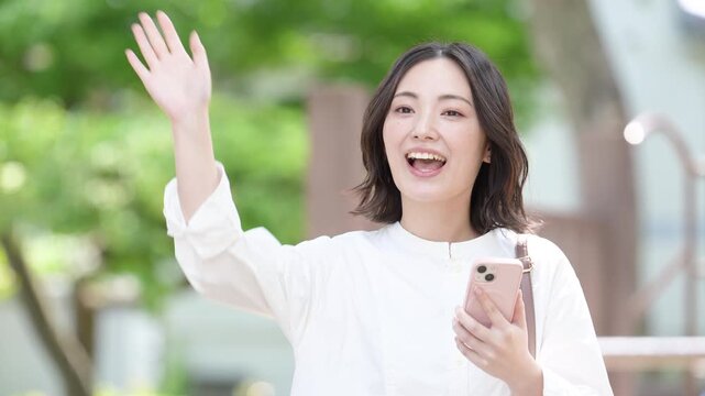 新緑の屋外でスマートフォンを持ちながらバイバイと手を振る女性の待ち合わせでのライフスタイルイメージの動画