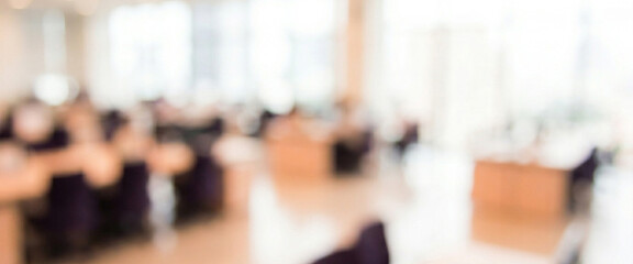 Fototapeta premium A blurred view of a spacious office environment with multiple desks, chairs, and large windows allowing natural light to enter the space.