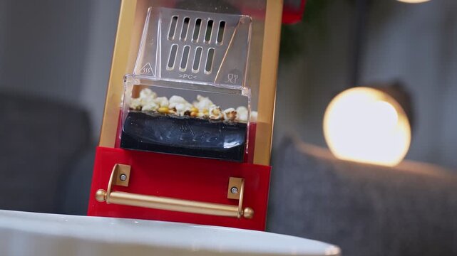 a vintage red and gold popcorn machine popping fresh corn kernels into a white bowl in a cozy home interior with warm bokeh lighting. Perfect for cinema and snack-themed content.