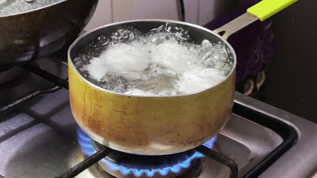 White eggs boiling in a small pot of water on a kitchen gas stove with blue flames. Bubbling hot water and steam during breakfast food preparation.