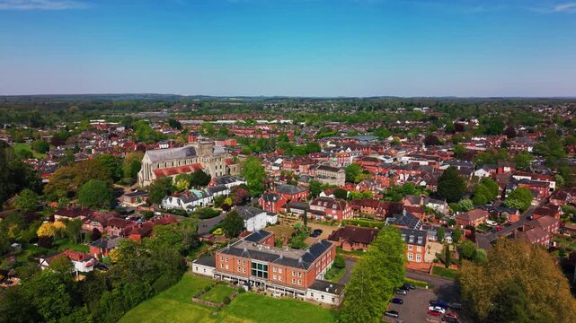 Wide aerial drone footage of Romsey Abbey and the surrounding Hampshire town skyline, showing historic rooftops, green spaces, trees, streets, residential areas, and countryside beyond on a sunny day.