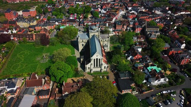 Top down aerial drone footage of Romsey Abbey in Hampshire, showing the historic abbey church, churchyard, surrounding rooftops, streets, green spaces, trees, and town centre on a sunny day.