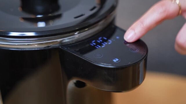 A close-up shot of a finger pressing the touch-sensitive digital display of a modern black electric kettle in a sunlit kitchen. The display shows 99 degrees on the sleek appliance panel.