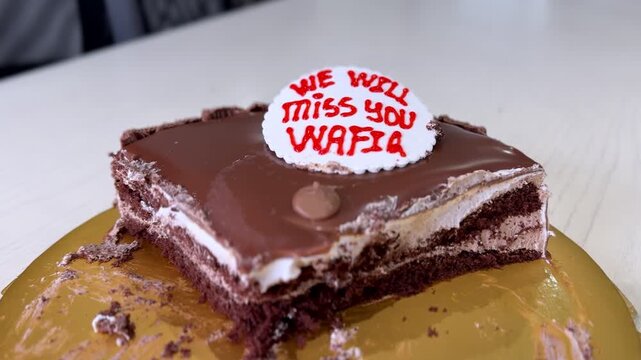 Close-up of a chocolate layered farewell cake with a message saying "We will miss you Wafia" on a gold base during a daytime office party. Cinematic shallow focus on the dessert detail.
