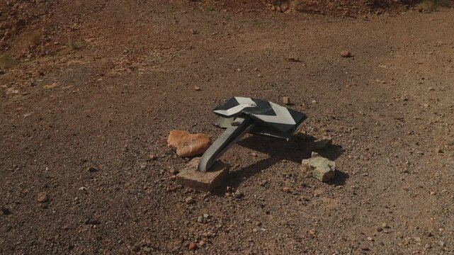 Toppled bent metal road sign with black and white arrow markings collapsed on dirt ground beside rocks in Moroccan desert