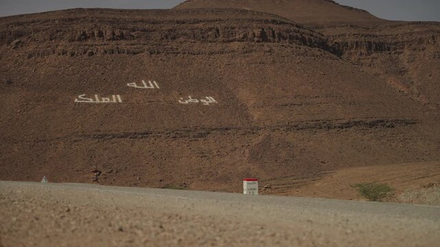 Moroccan national motto in Arabic painted across rocky hillside along desert road